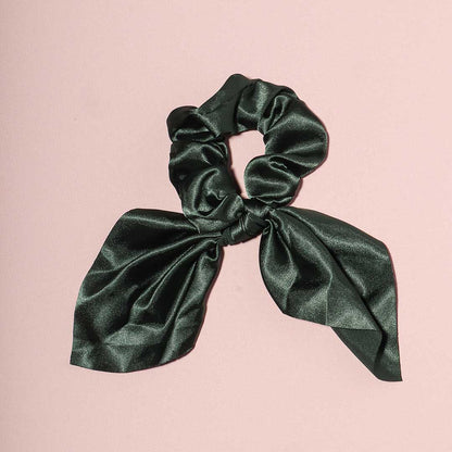 Elle dark green satin scrunchie with a chic bow attachment, displayed flat on a light pink background.