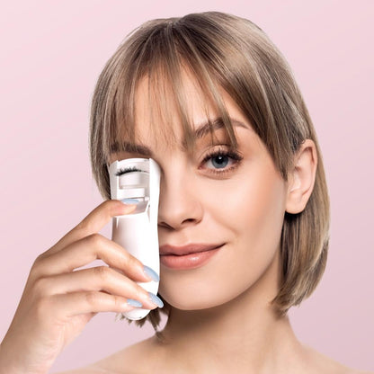 Model using white Tasha Heated Eyelash Curler to lift and curl lashes for a voluminous look against a pink background.