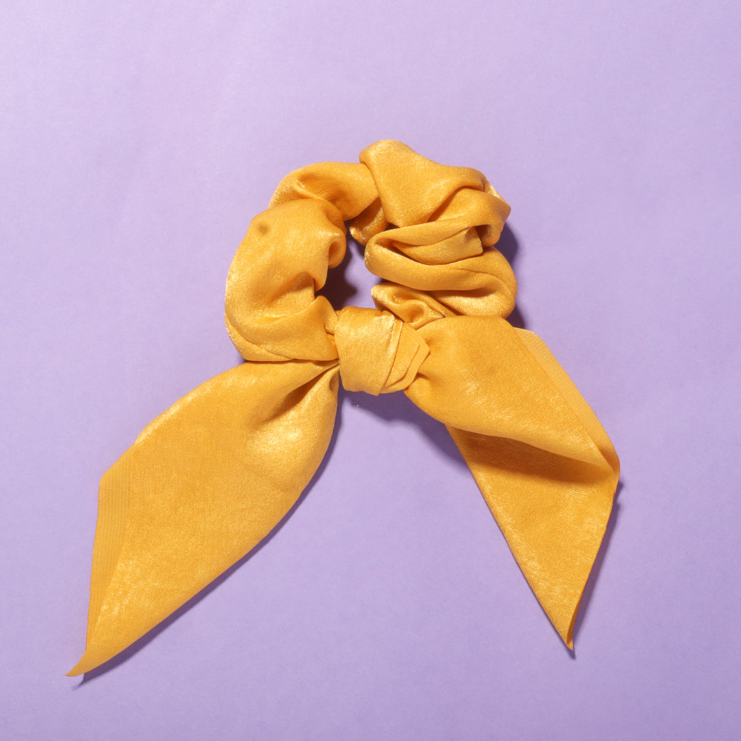 Yellow Elle satin scrunchie with chic bow detail on a purple background.